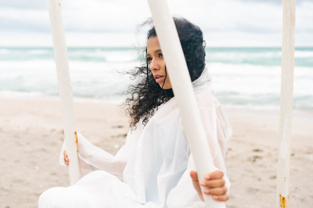 Woman with curly hair in white clothing, sitting on a swing by a windy beach.